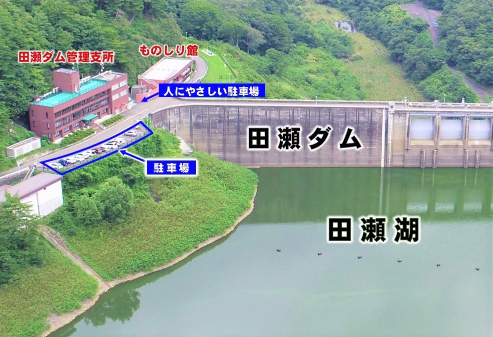 田瀬ダムを上空からうつした画像。左側に管理支所と駐車場、右側にダムの堤体とダム湖がある。