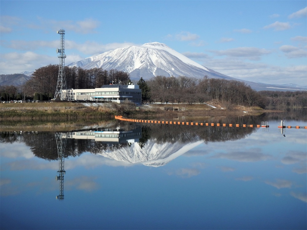 雪をかぶった岩手山と青空、白い雲が湖面に鏡のように写っている。